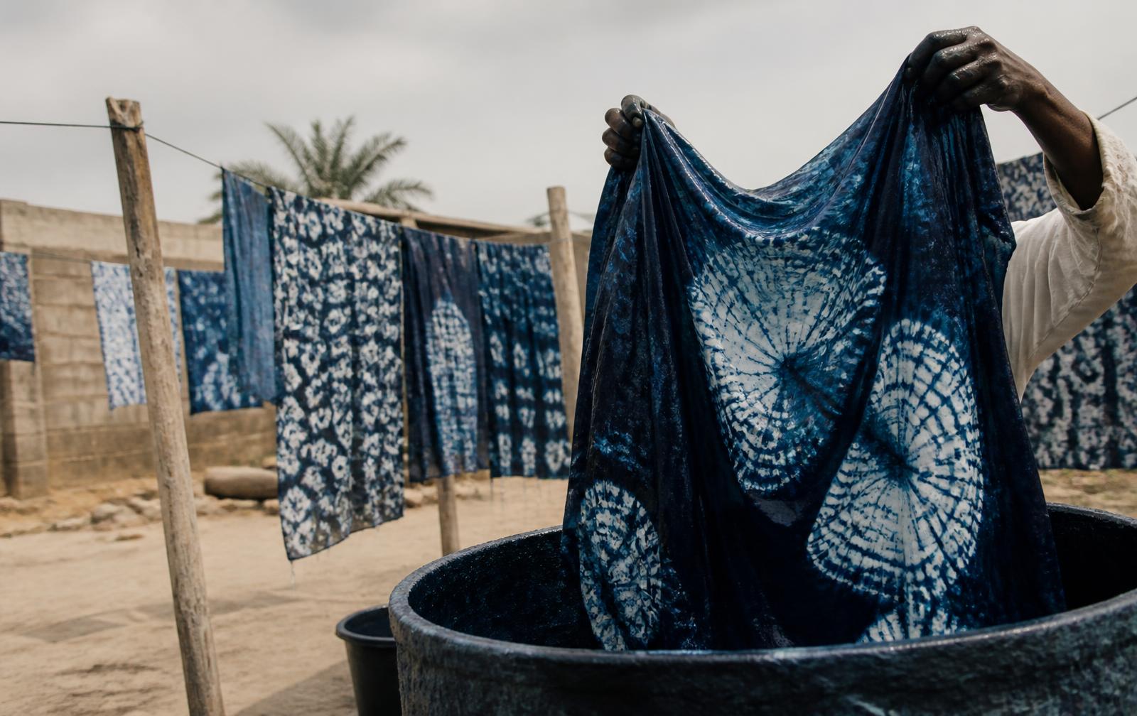 Hands lifting freshly indigo-dyed adire cloth from a dye vat in a Lagos dye yard, patterned cloth drying behind